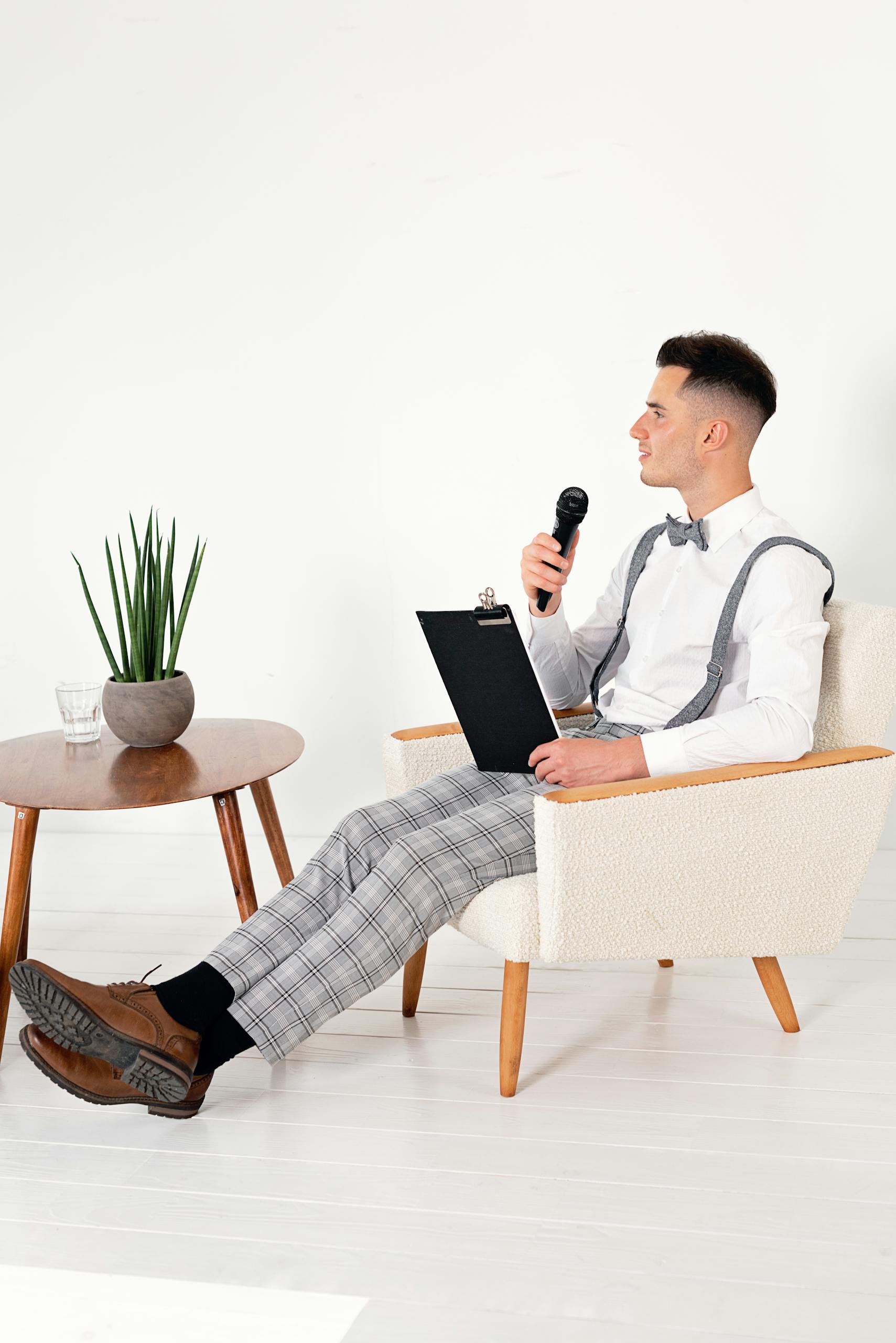 Young man in white shirt and suspenders holding a microphone and clipboard while sitting in a stylish chair indoors.